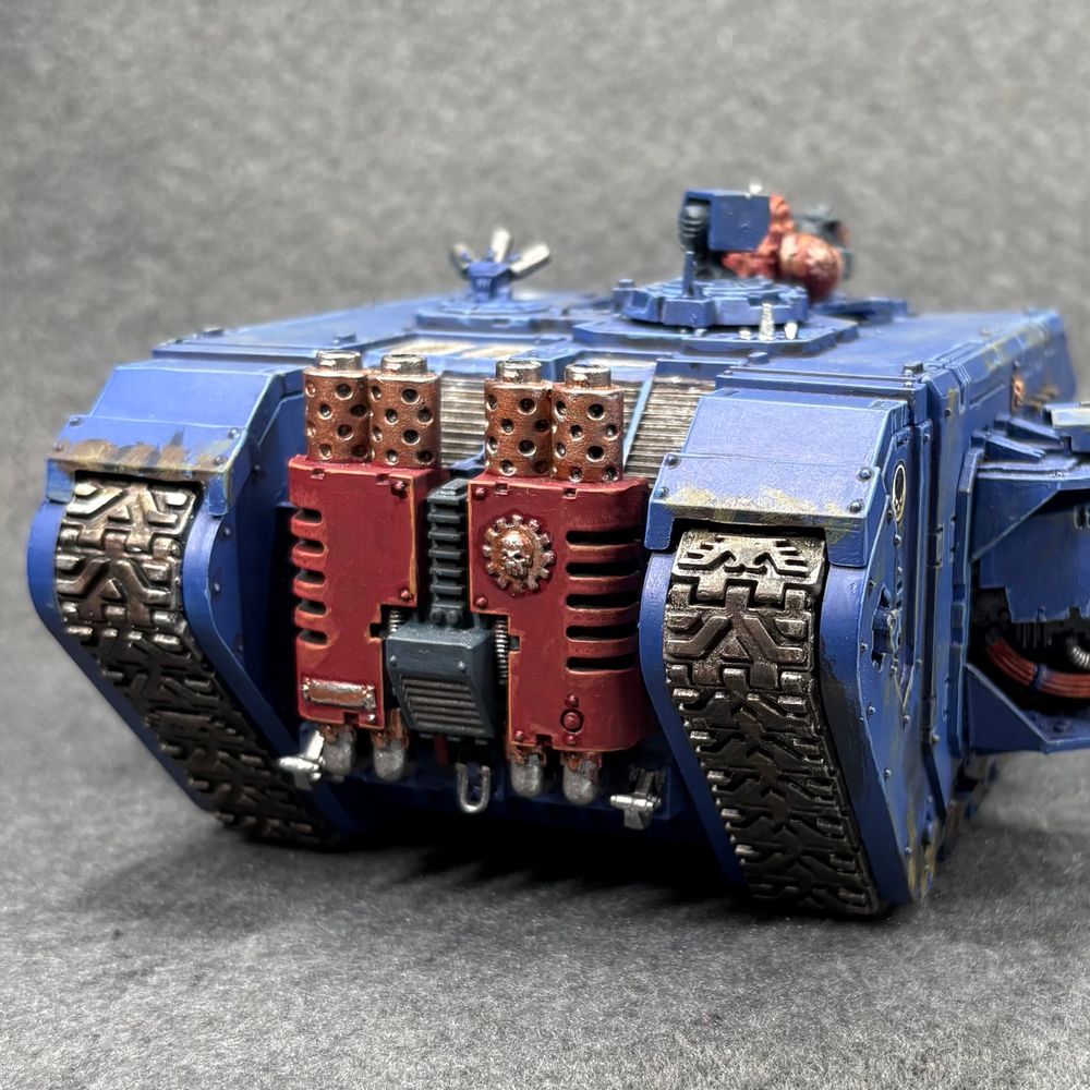 Rear view of a large boxy tank painted ultramarine blue with a red engine compartment. It has large tracks, sponsons with laser cannons, a marine popped out the top with a pintle-mounted gun, and lots of road grime. 