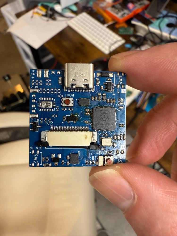 A hand holding a blue printed circuit board which has an RP2040 installed with various connectors for various peripherals.