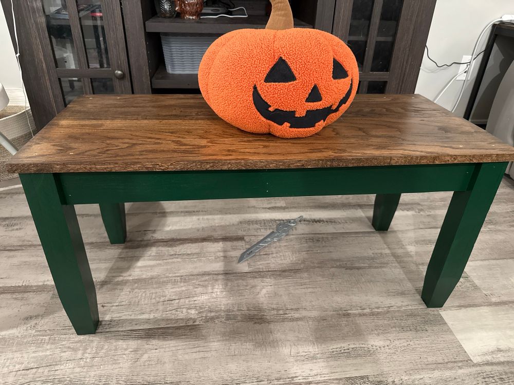 A long and slender coffee table with a dark oak top and a green base. Atop the table is a jack-o-lantern pillow with a big smile.