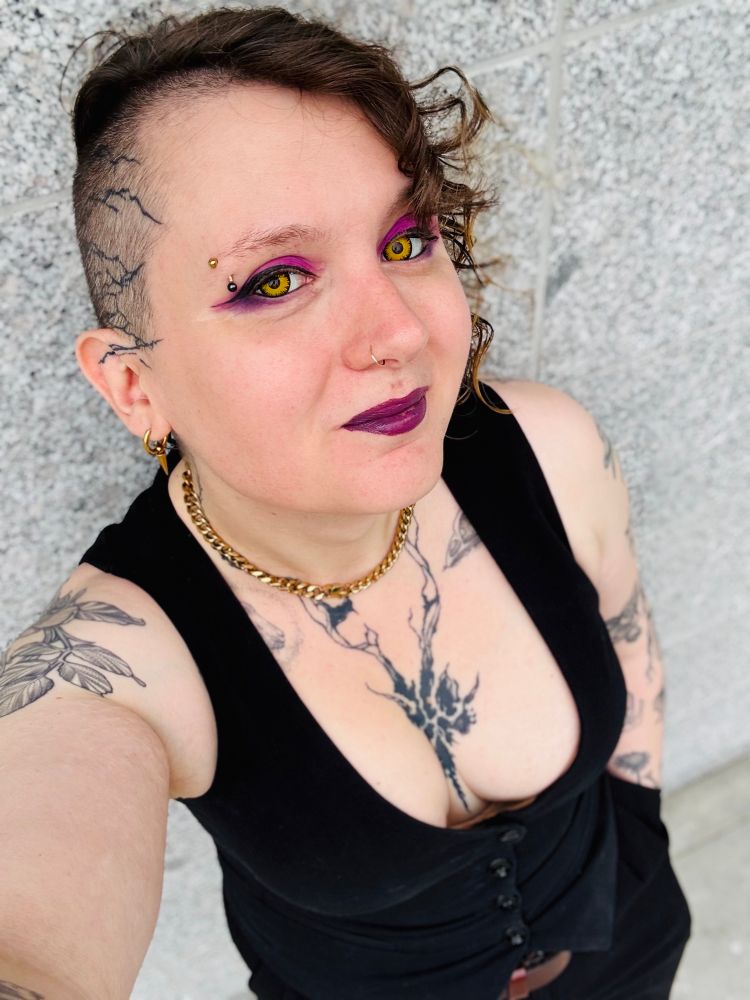 Andrea wearing a waistcoat with heavy cleavage, as well as purple/pink makeup and yellow contacts
