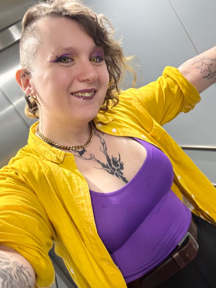 Andrea dressed in purple, yellow, and black (and the white of the non binary flag is represented by her white-ass skin)