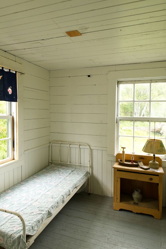 A sparse bedroom interior, with a simple single bed, a side table with a lamp and a couple of small objects, all lit by two windows.