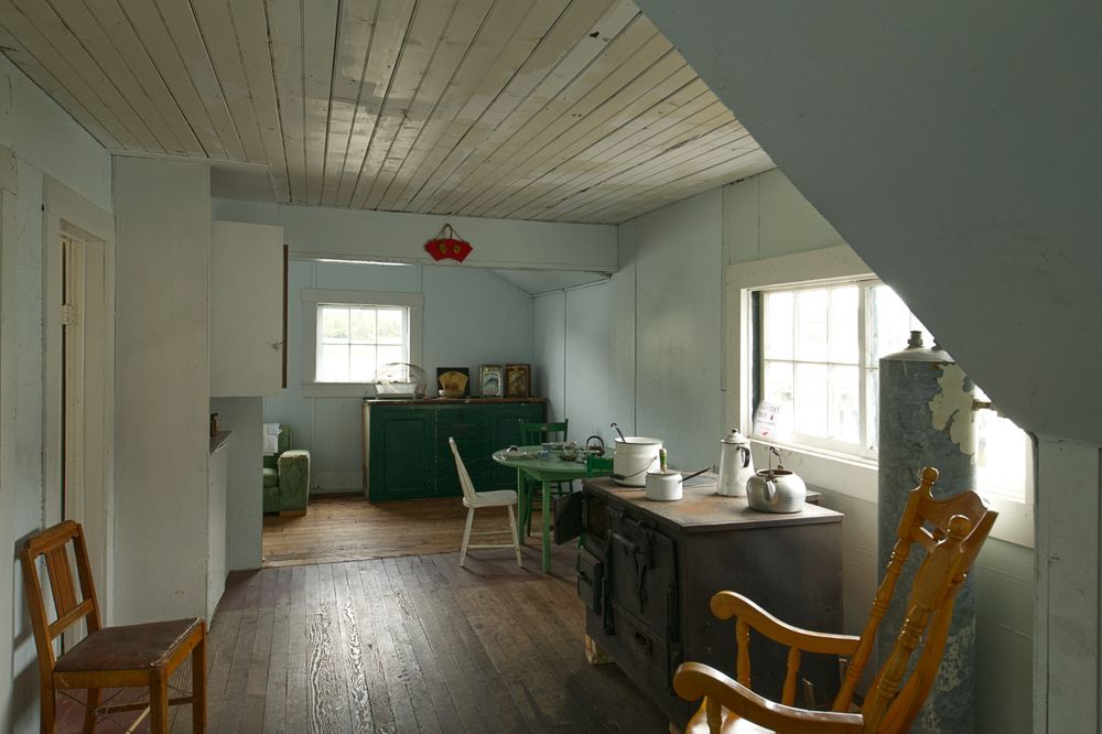 A simple wooden domestic space, with a woodstove, several wooden chairs, and a modest wooden dining table.