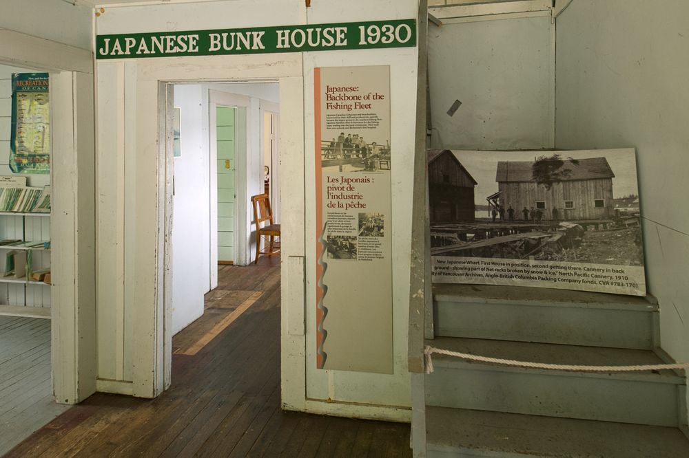 A very modest wooden domestic interior, evidently a foyer, with doors leading to other rooms and stairs leading up. Modern signage identifies it as the 'Japanese Bunkhouse'