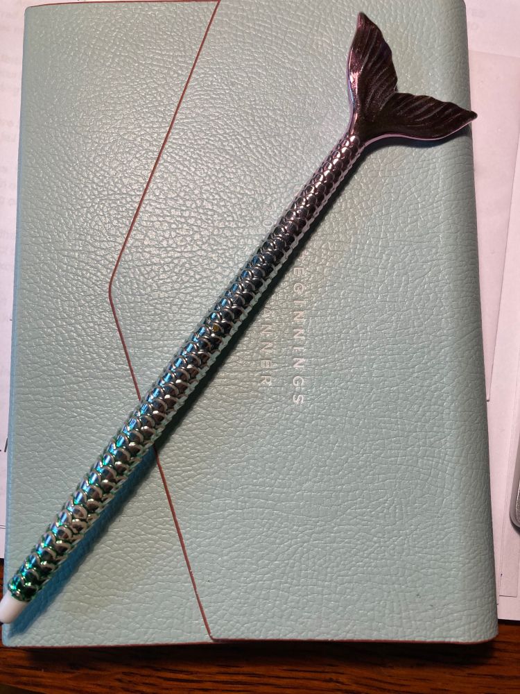 A ball point pen designed to look like a mermaid tail, with metallic aqua and pink scale texturing and a tail at the end. 