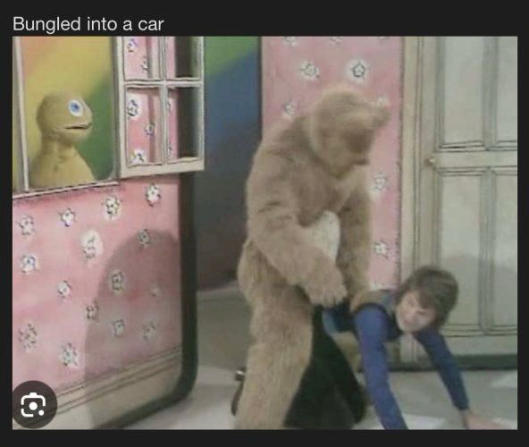 Still from 1980’s Uk childrens tv show “Rainbow”. The male presenter (Geoffrey) is knelt on the floor, while a character in a bear suit (Bungle) is *probably* helping him up but is stood behind and astride Geoffrey. A hand puppet with a zip for a mouth is watching from an open window on the left side of the frame