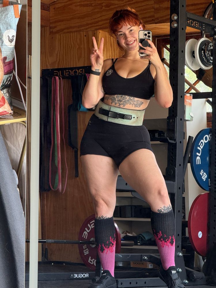 Mirror selfie of Siri in the garage gym wearing a black sports bra and shorts, lifting belt, deadlift slippers and socks, and a bar loaded with 100kg behind her 😤💪🏼