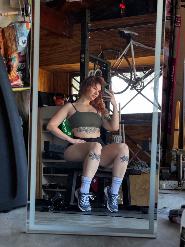 Siri taking a mirror selfie in the garage gym while sitting on a weight bench and looking VERY cute damnit