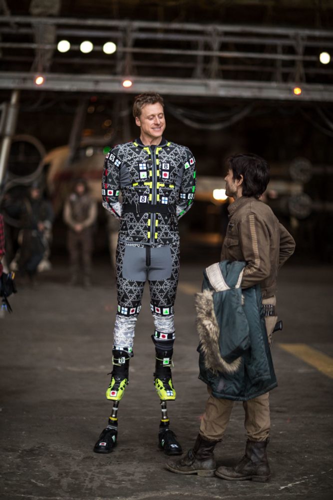 Behind the scenes photo from Andor of Alan Tudyk, wearing a motion capture suit and standing on stilts, talking to Diego Luna
