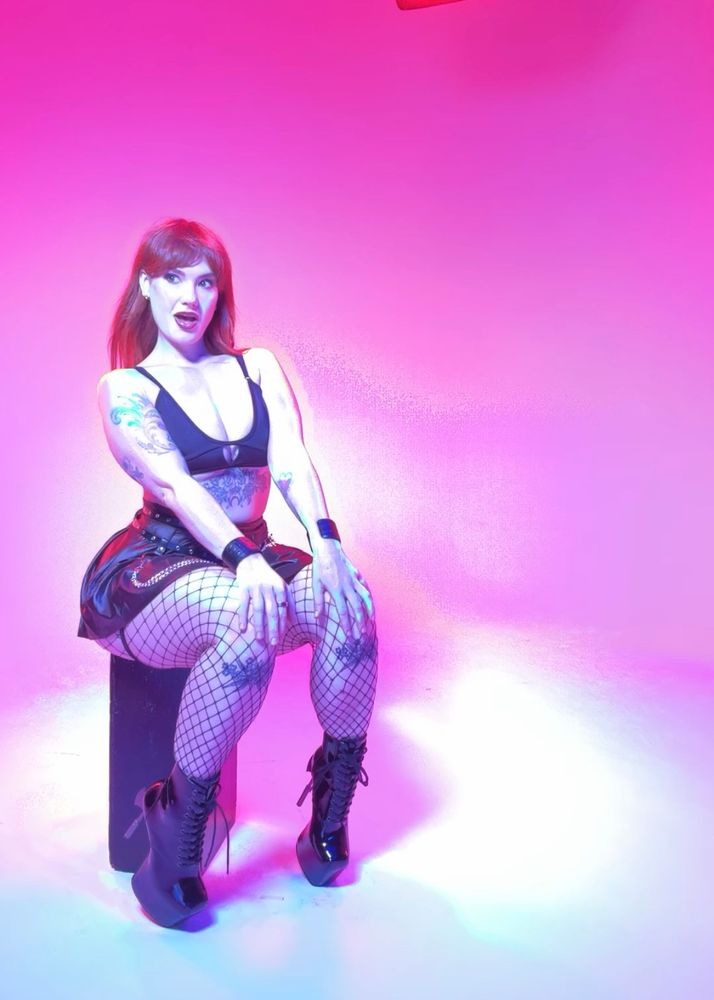 [a very artsy high contrast photo] Siri striking a pinup-style seated pose on an apple box, wearing an alt goth style black outfit with fishnets and shiny stiletto boots, black skirt and crop top, against a white backdrop with pink lighting 