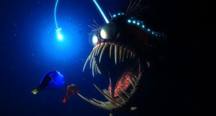 A still from the movie "Finding Nemo", where two fish (the orange Nemo and blue Dory) are wide-eyed, swimming in the dark ocean up to a glowing ball of light. On the right side of the frame, you can see that the light is attached to the antennae of an angler fish, which emerges from the darkness with a wide-open mouth full of enormous sharp teeth.