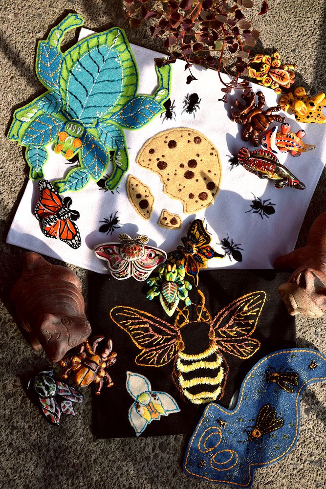 A picture of some of my stuff, there are a pair of embroidered napkins (one white with some ants around a cookie, and a black one with an orchid bee). There are also a pair of embroidered patches (one of a leaf bug and another one of a planaria with a firefly motiff). And a bunch moth, beetle and other creature polymer clay pins