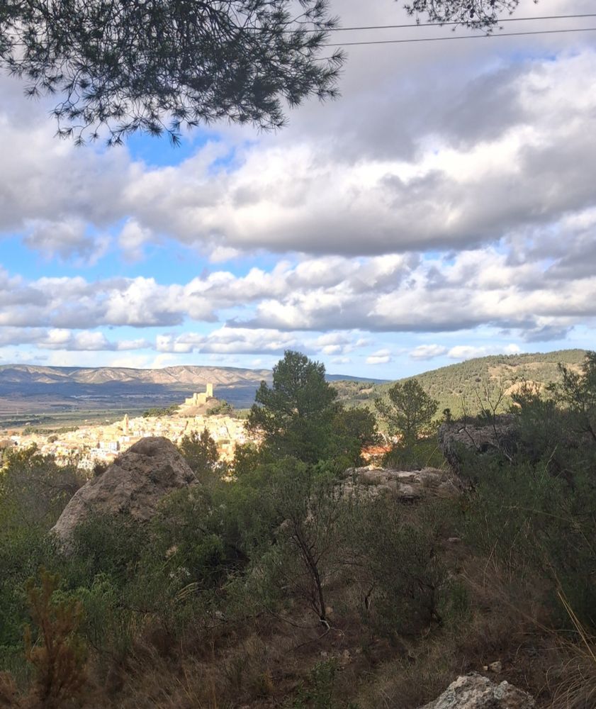 Biar, Alicante,  village and castle, sunlit in the background,  blue sky teeming with cumulonimbus, pine scrub and rocky crags in foreground.