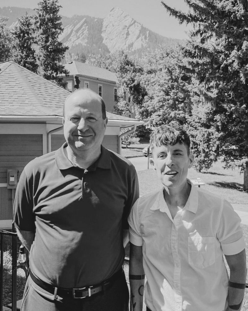 A black and white photo featuring Governor Jared Polis standing outdoors alongside Poet Laureate Andrea Gibson.