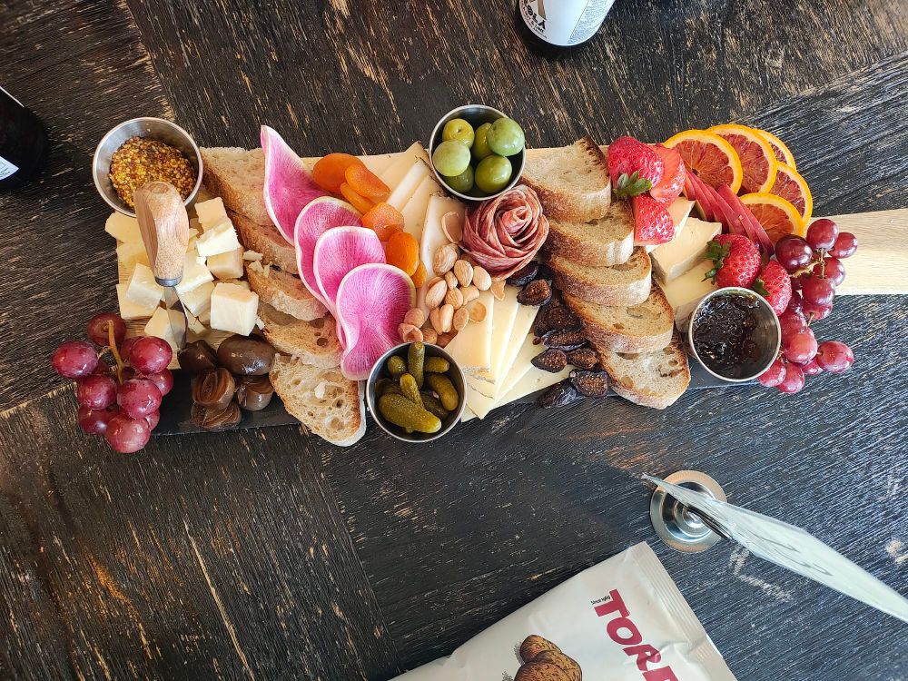 Photo of a Charcuterie board, four cheeses, meat, olives, figs, bread, cremini onions, watermelon radish, red grapes, blood orange,figs, gala apples, marcona almonds, dried apricots, cornichons, strawberries, mustard, and fig marmalade.