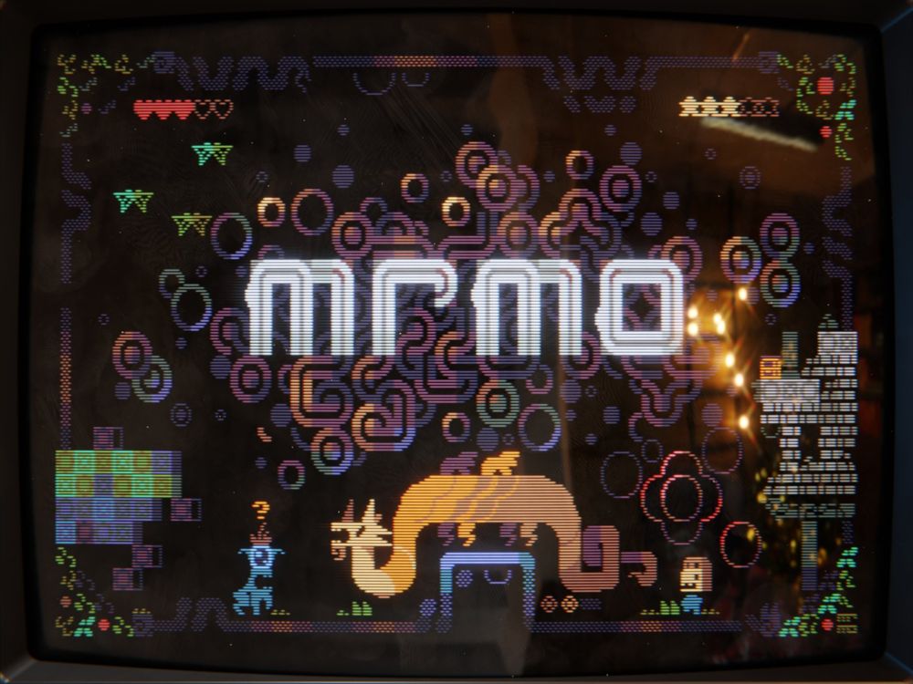 A 3d render of a scene that depicts a vintage CRT display, showing a colorful textmode art, mostly abstract, with several creatures including an orange dragon. The big, white letters in the middle spell out MRMO.