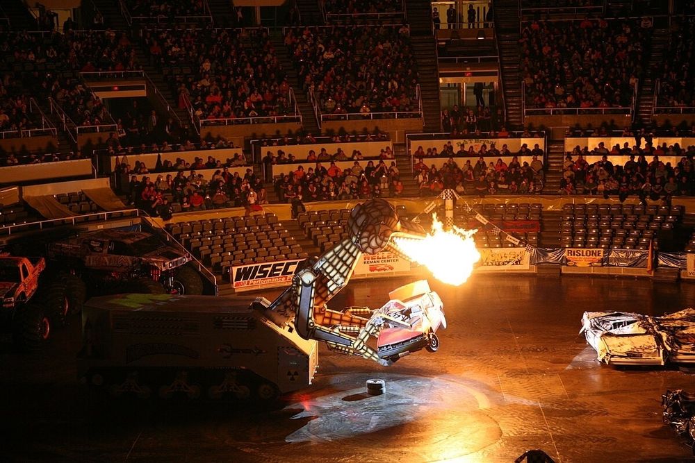 A still from a monster truck event where a large firebreathing robot dragon thing is holding a wrecked car in its claws