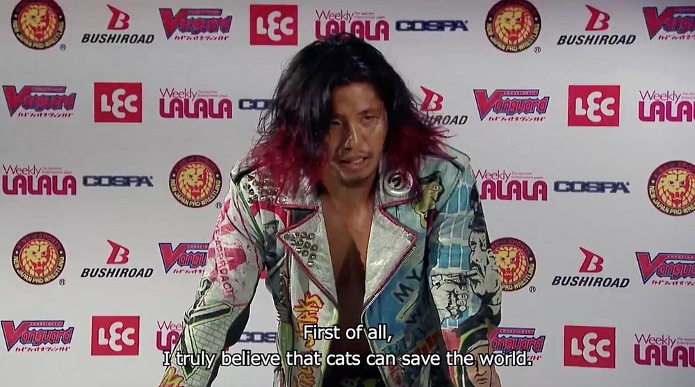 Still from a NJPW press conference showing Hiromu Takahashi speaking with text that reads "first of all, I truly believe that cats can save the world"