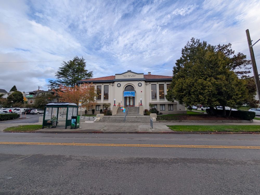 A Carnegie style library with a blue banner that reads "Welcome back to your Library!" There are blue balloons and a red ribbon that cross the steps. At the street is a covered bus stop. This photo is further back with a blue sky covered with feathery clouds.