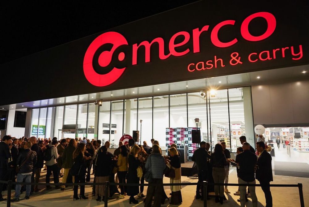 A night-time shot of a queue waiting outside a Comerco cash and carry. The store logo is lit up in red.
