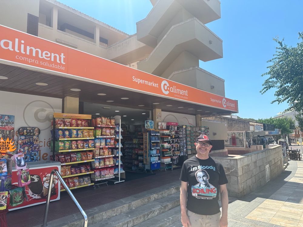 A smiling buffoon in front of a small supermarket shop front. The orange and white sign reads: Supermarket Coaliment