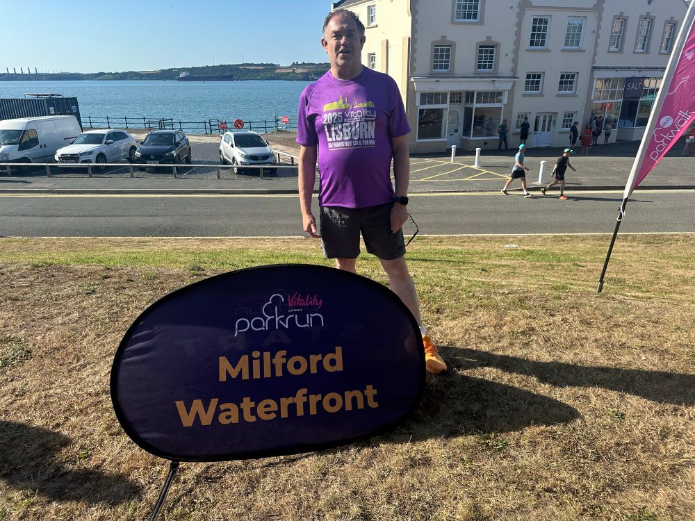 Me standing behind a parkrun Milford Waterfront sign, with Milford Marina and the estuary in the distance