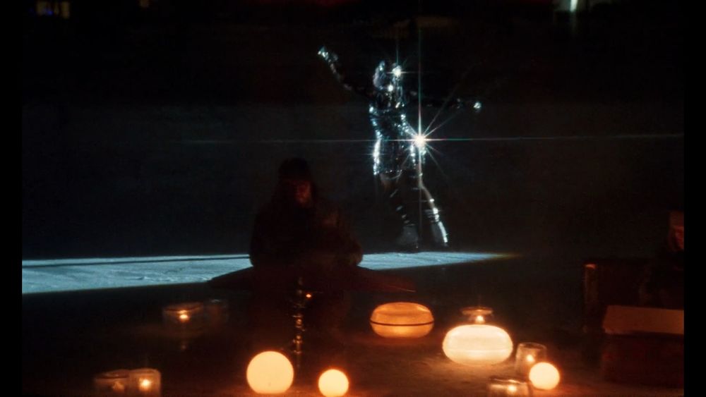 Still from Universal Language; a woman in a glittering spangled dress ice skates in a spotlight; in the foreground, two men surrounded by soft yellow lamps on the ice play instruments.