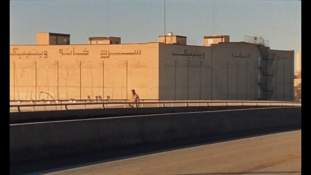 Still from Universal Language; wide shot of a man crossing an otherwise empty concrete bridge or highway, with a windowless building lit in golden dusky light in the background, with Arabic lettering on it.