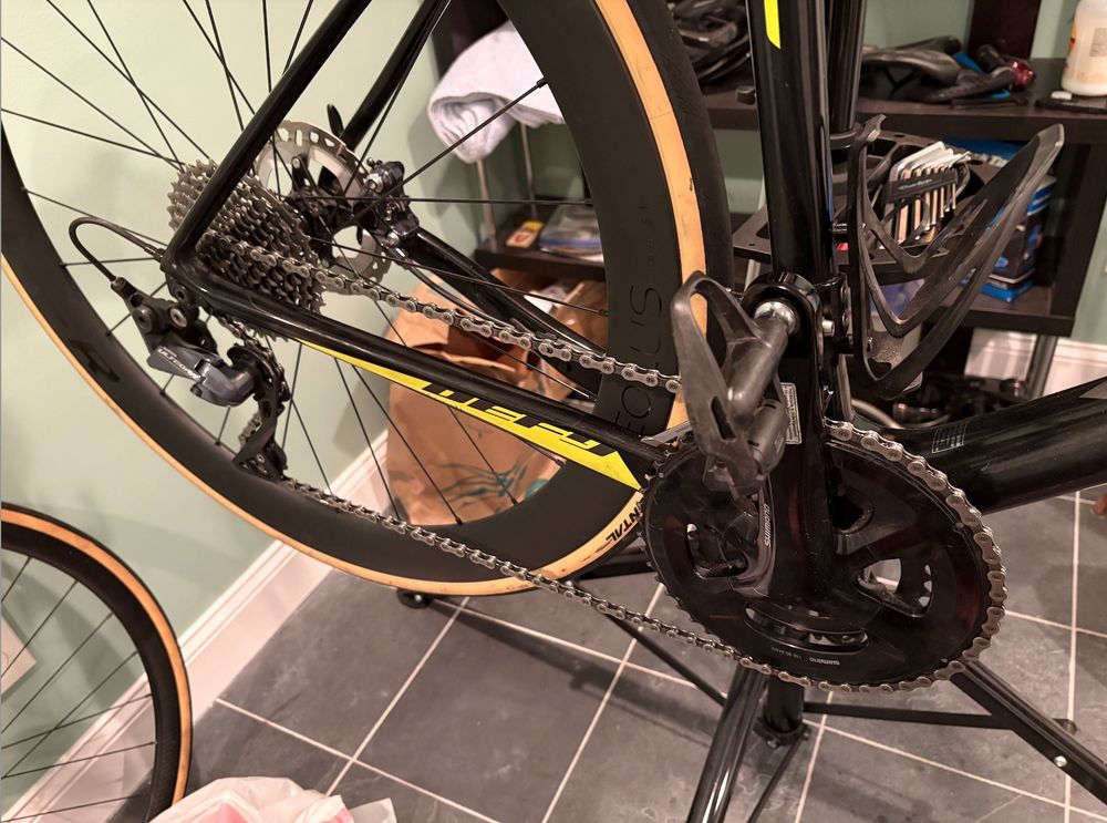 Image of the rear wheel, gears, and cranks of a road bike lifted off the ground on a work stand. Various equipment is in the background.