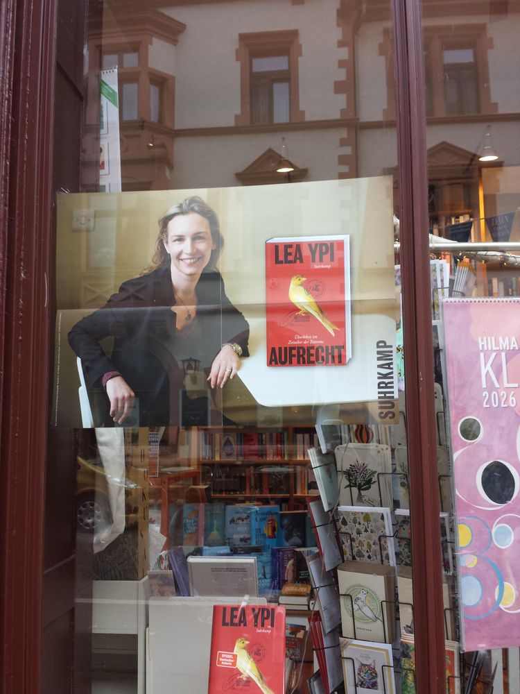 Ein kleiner Buchladen. Die Schaufenster sind individuell gestaltet. Die Auswahl an Büchern klingt interessant und unterscheidet sich vom Bahnhofsladen. Im Fenster spiegelt sich eine Altbaufassade, mit Farben, die zum Herbst passen.