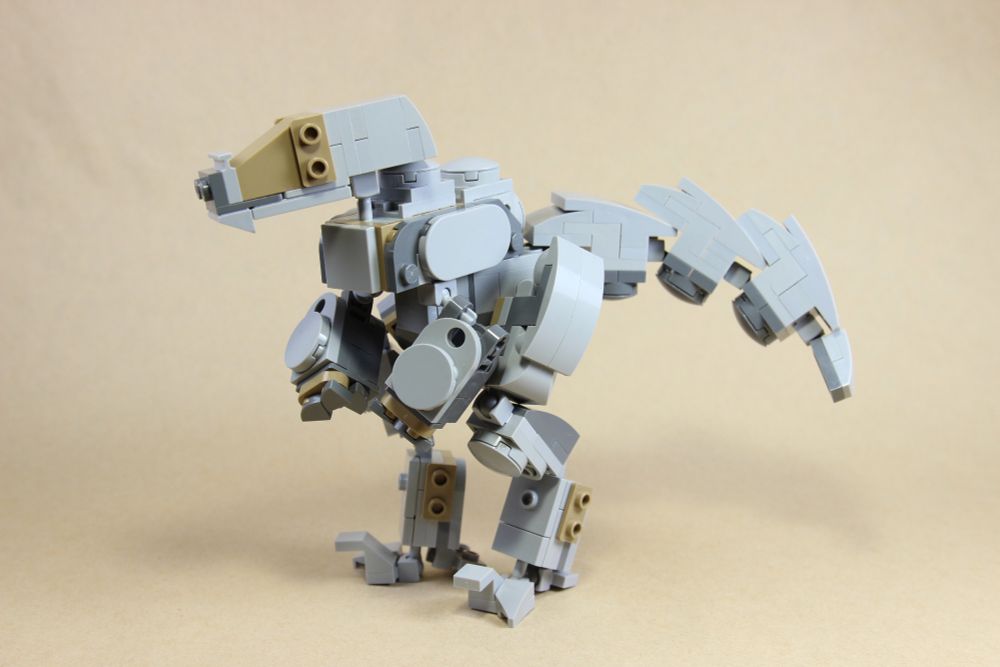 Lego model. A grey robotic velociraptor-type dinosaur. It has some dark tan accents on its face and limbs, a big segmented tail, and four eyes. Side view, slightly to the front.