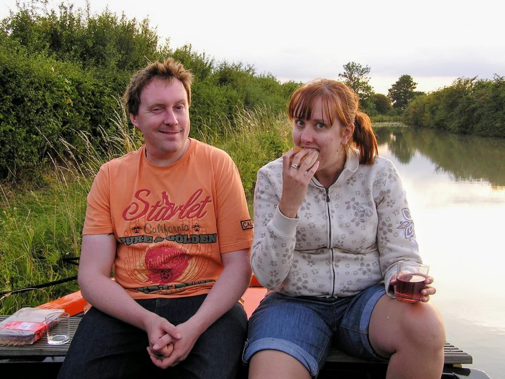 A man and a woman sit beside a calm canal with greenery on both sides. The man, wearing an orange t-shirt with printed text, looks at the camera with a playful expression. The woman, wearing a light patterned hoodie and denim shorts, is eating a sandwich and holding a drink. A small container of food rests on the bench beside them, and the evening light reflects off the water behind them.