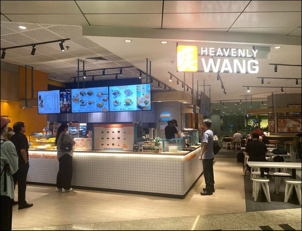 A bright cafe counter with a lit sign that reads Heavenly Wang. Several customers stand in front of the counter looking at digital menu boards. Staff prepare food behind the counter. Tables and seated diners are visible in the background.