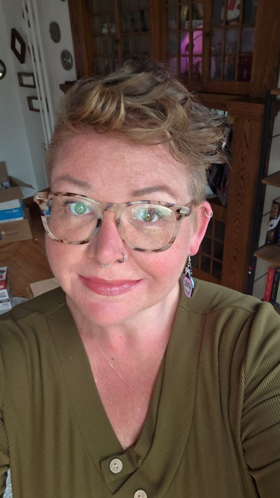 a selfie of a smiling nonbinary person, they have short curly hair, tortoiseshell glasses, and black earrings that look like little bottles of poison. they are alao wearing an olive green v neck sweater with some wooden buttons at the neck 