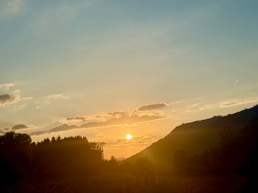 Landschaftsfoto, das einen Sonnenuntergang zeigt. Die Sonne befindet sich tief am Horizont und ist teilweise von dünnen Wolken verdeckt. Im Vordergrund sind dunkle Silhouetten von Hügeln und Bäumen zu erkennen, während der Himmel einen Übergang von warmen Gelb- und Orangetönen zu kühleren Blau- und Violetttönen aufweist.

 

