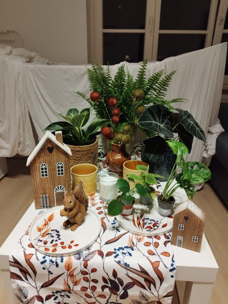 einige zimmerpflanzen, holzhäuschen, eine eichhörnchen figur und eine kerze auf einem tisch mit läufer mit blattmotiven