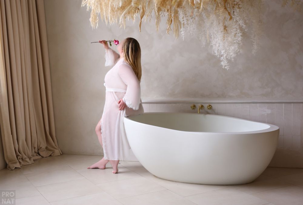 White, plus-size woman wearing a long, sheer white robe holding and smelling a single rose while standing next to a free-standing tub.