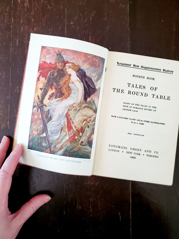 A hand holds open a book. On the left page is a colour plate showing Lancelot and Guinevere. On the right page is the title page of the book: Tales of the Round Table.