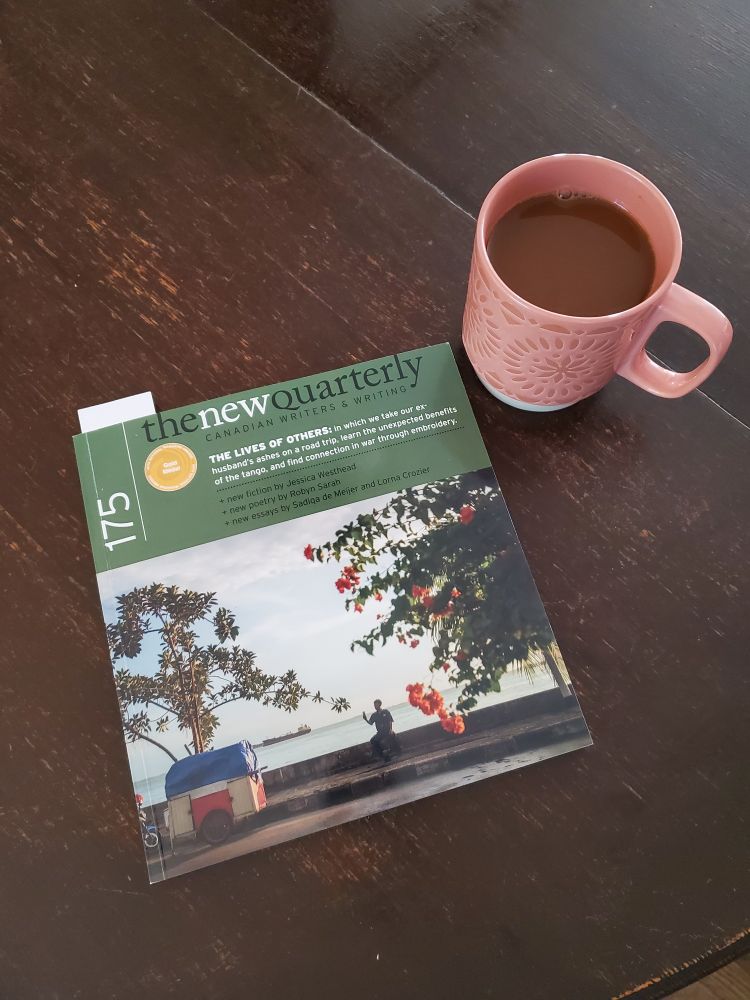 A periodical called The New Quarterly, featuring a photo of a person sitting on a wall under some trees on the cover, lays on a table. A pink mug filled with coffee sits beside it.