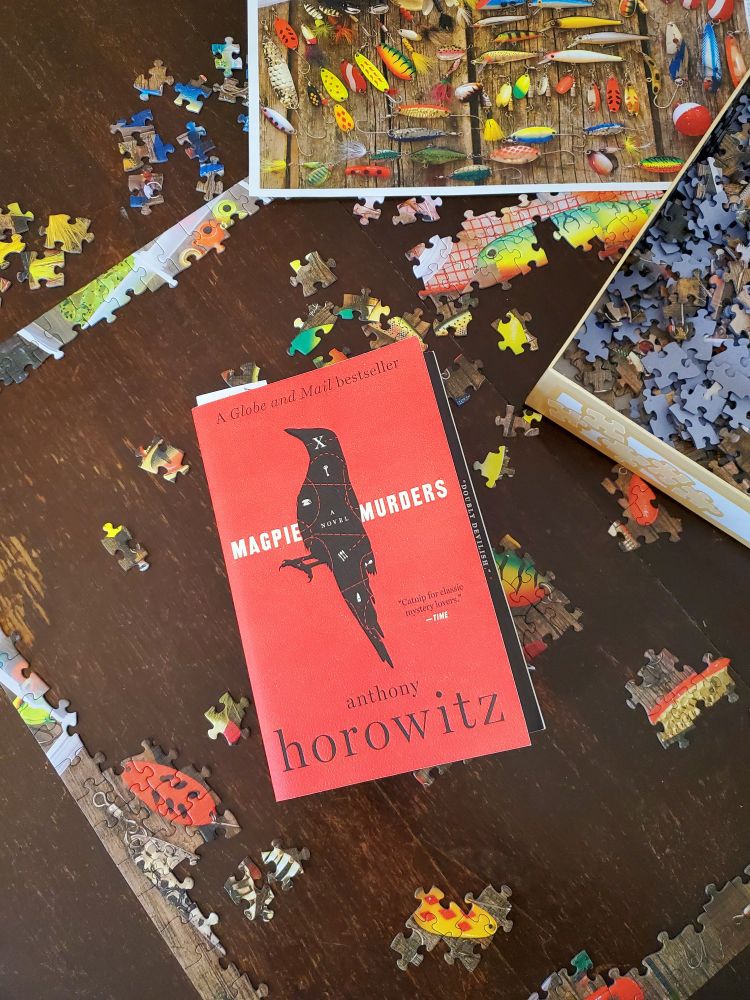 A book with a red cover, entitled "Magpie Murders" by Anthony Horowitz, sits atop a table with a partially completed jigsaw puzzle