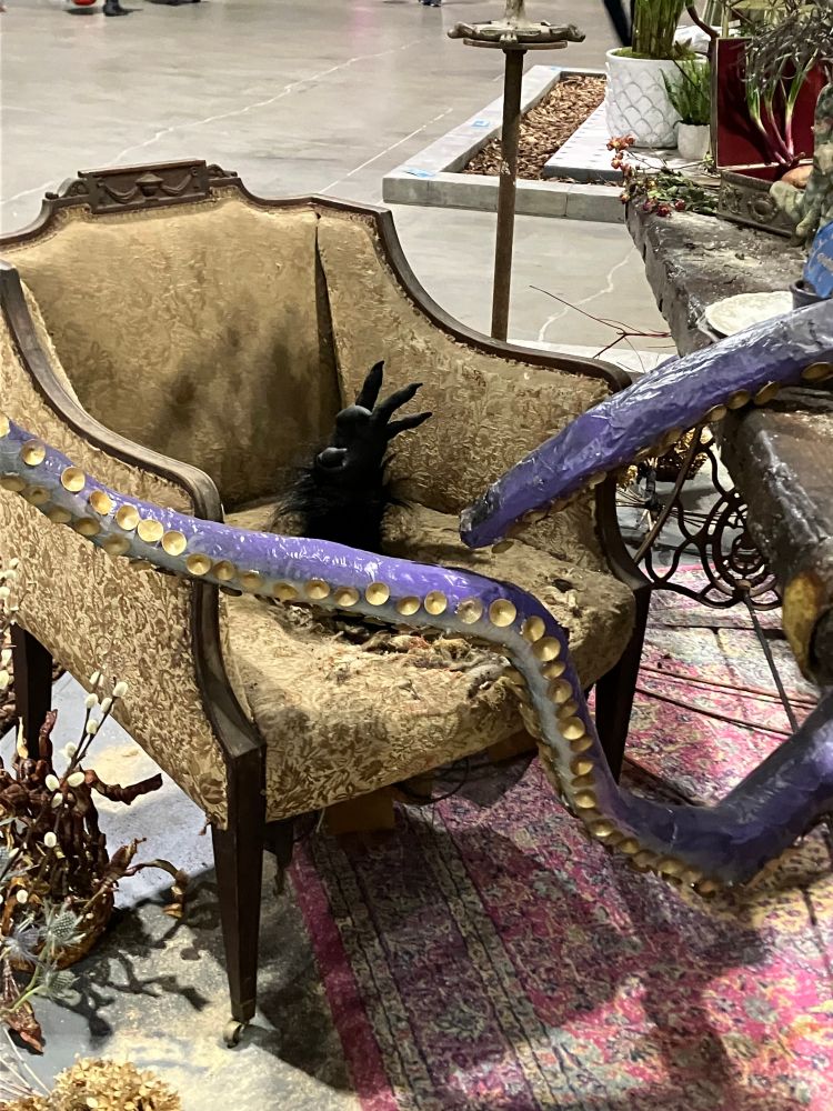 Close up detail of "Of Gods and Monsters," a display by the Grumblethorpe Youth Volunteers at the 2025 Philadelphia Flower Show. A monstrous, furred, black arm reaches its clawed hand extradimensionally through the seat of H.P. Lovecraft's seat at the imagined table he might have shared with Mary Shelley.