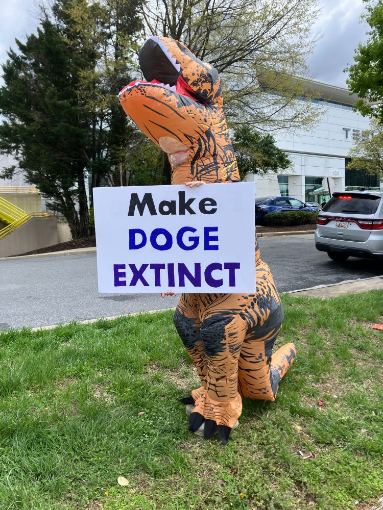 Person in inflatable tyrannosaurus costume holding sign saying “Make DOGE Extinct”