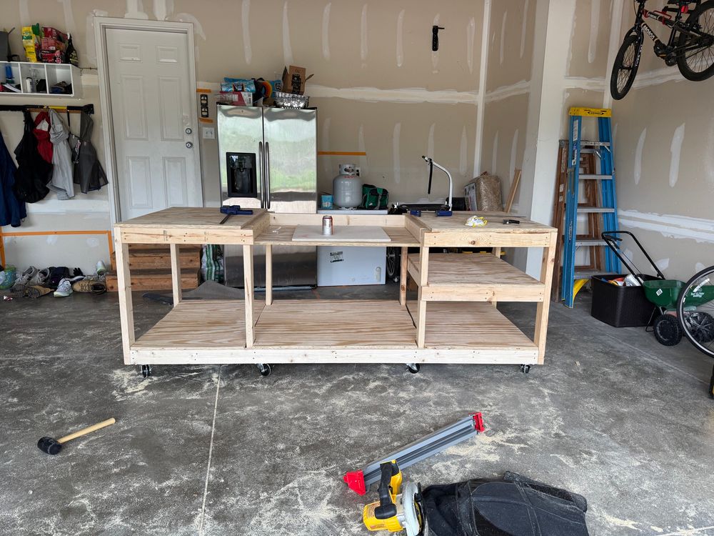 A finished, rolling workbench in a garage. The whole workbench is approximately 8ft long, 3.5 ft deep, and about 40 inches tall.  The middle section of the workbench is slightly lower than the tabletops on either side to accommodate a miter saw. There is saw dust everywhere, and some tools in the foreground. There is a cold seltzer on the workbench. 