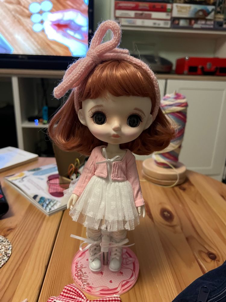 A Strawberry Fields Mori Lolli doll on a red and pink doll stand wearing a white dress with lace socks, white boots, and a pink cardigan and knitted hair bow