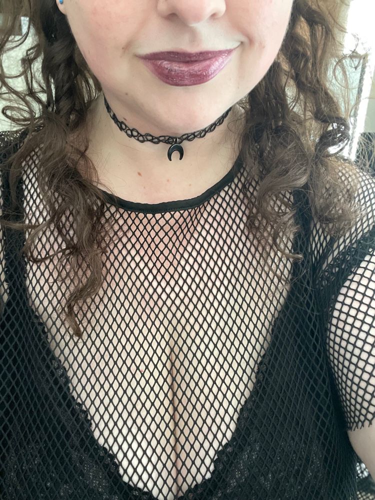 picture of phi in real life with a black bralette and fishnet top, a stretchy necklace with an upside down moon crescent, and dark glittery lipstick, with brown curls on either side of her face that cuts off at the tip of the nose
