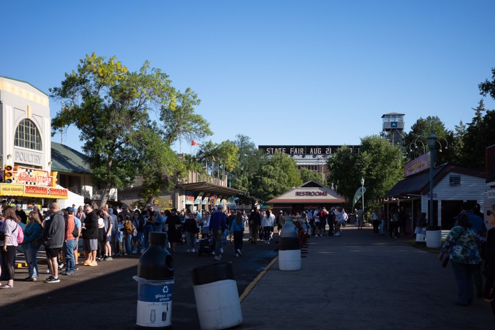 A bustling scene at a state fair showing crowds of people walking along a main thoroughfare lined with food vendors and buildings. In the background, a prominent sign reads 'STATE FAIR AUG 21' spanning across the pathway. On the left, there's a white building with 'POULTRY' signage and various food stands including one advertising 'Cinnamon Steak.' On the right side, a restrooms building is visible among other fair structures. The scene is set under a clear blue sky with mature trees throughout the fairgrounds, and recycling bins are placed along the walkway. The atmosphere appears lively with fairgoers of all ages enjoying the event.