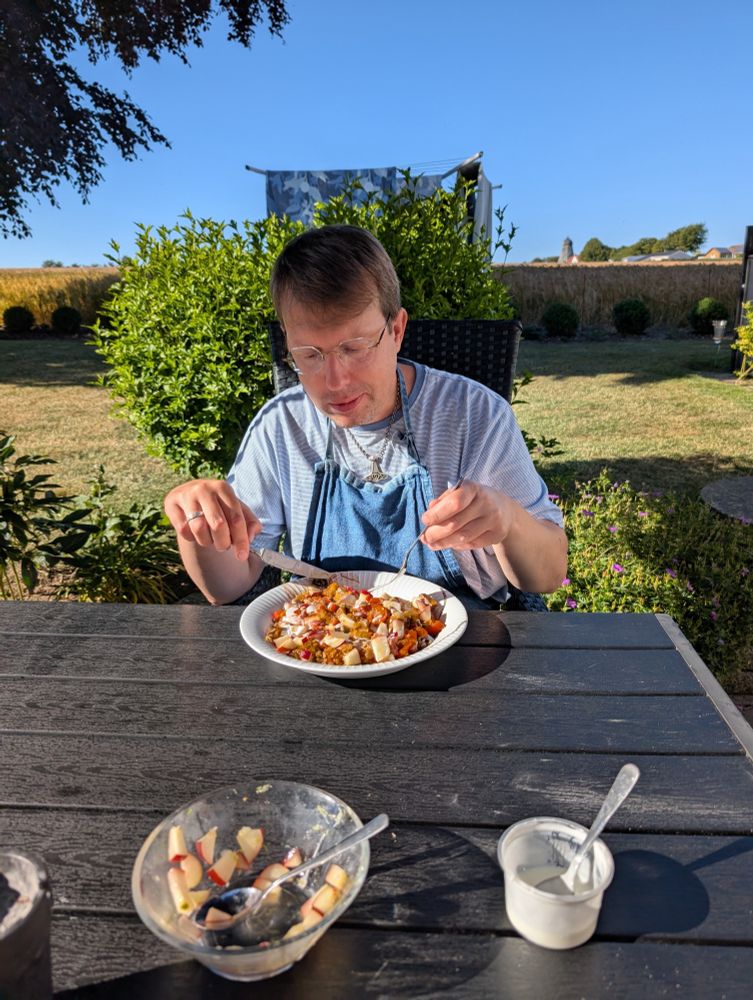 Dennis sidder ved havebordet på terrassen og spiser sin aftensmad. Han har en blå tshirt og et forklæde på. I Forgrunden på bordet står en skål med limenarinerede nektariner og en bøtte yoghurt.