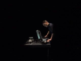 Man (Thomas Ankersmit) standing, performing on a modular synthesizer, against a black background. Photo credit: Udo Siegfiedt