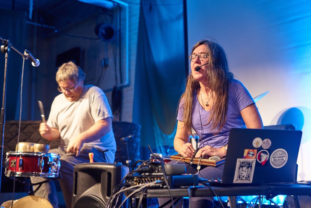 Photo of Chris Strunk performing on a single snare drum with upside down Tibetan bowl and table knives. Andrea Pensado is singing in the foreground, performing with an electronic instrument in her lap.