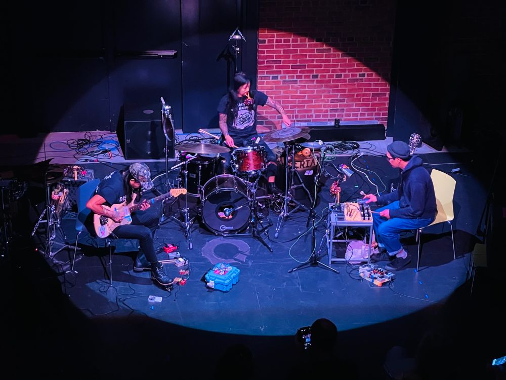Photo of MANAS (Tashi Dorji, guitar; Thom Nguyen, drums; Che Chen, percussion) performing (lit by a spotlight) at the Cambridge Foundry. Shot from the lighting booth. Photo by Joseph Lark-Riley